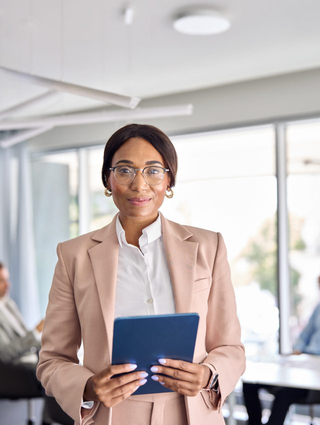 Confident African American business woman manager standing at office team meeting. Portrait of elegant smiling professional female company executive leader holding digital tab in board room. Vertical.
